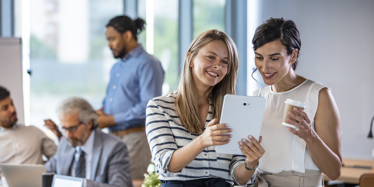 Young colleagues coworkers working together in company meeting room using digital tablet. Smiling businesswomen using digital tablet. Female business professionals are working on project. They are wearing formals in office.